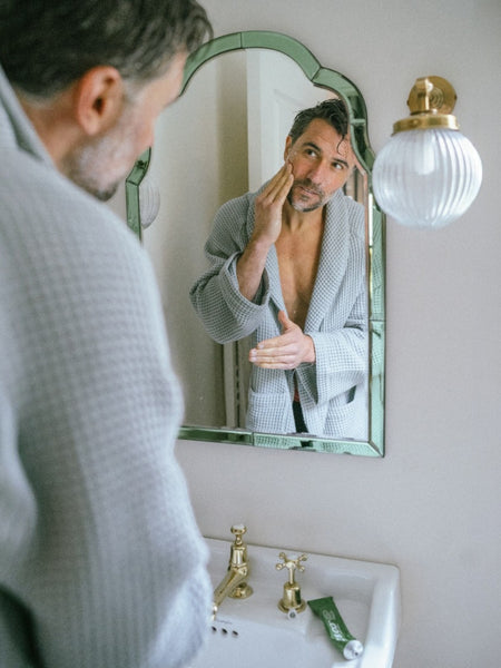 Baz & Co Skincare man in grey robe applying brightening exfoliator and cleanser to face in bathroom.