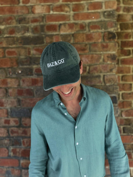 Baz & Co Skincare dark green washed cotton baseball cap with embroidered logo, worn by a man smiling against a brick wall.